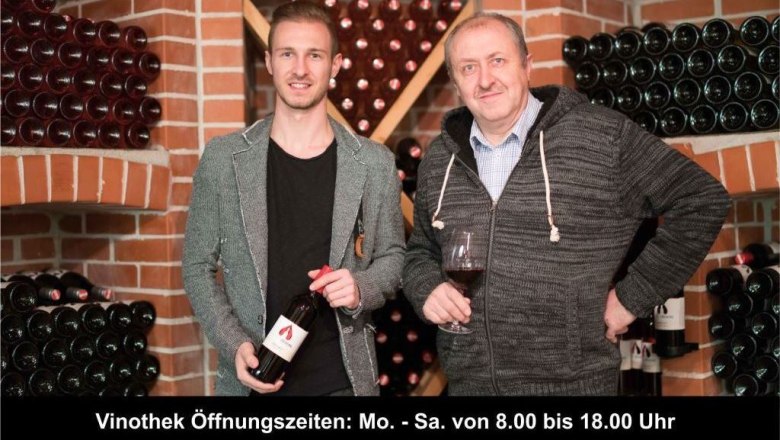 Two men in a wine cellar, surrounded by bottles of wine, one holding a glass of wine.