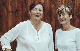 Zwei Frauen in weißen Blusen stehen vor einer Holzwand und lächeln.