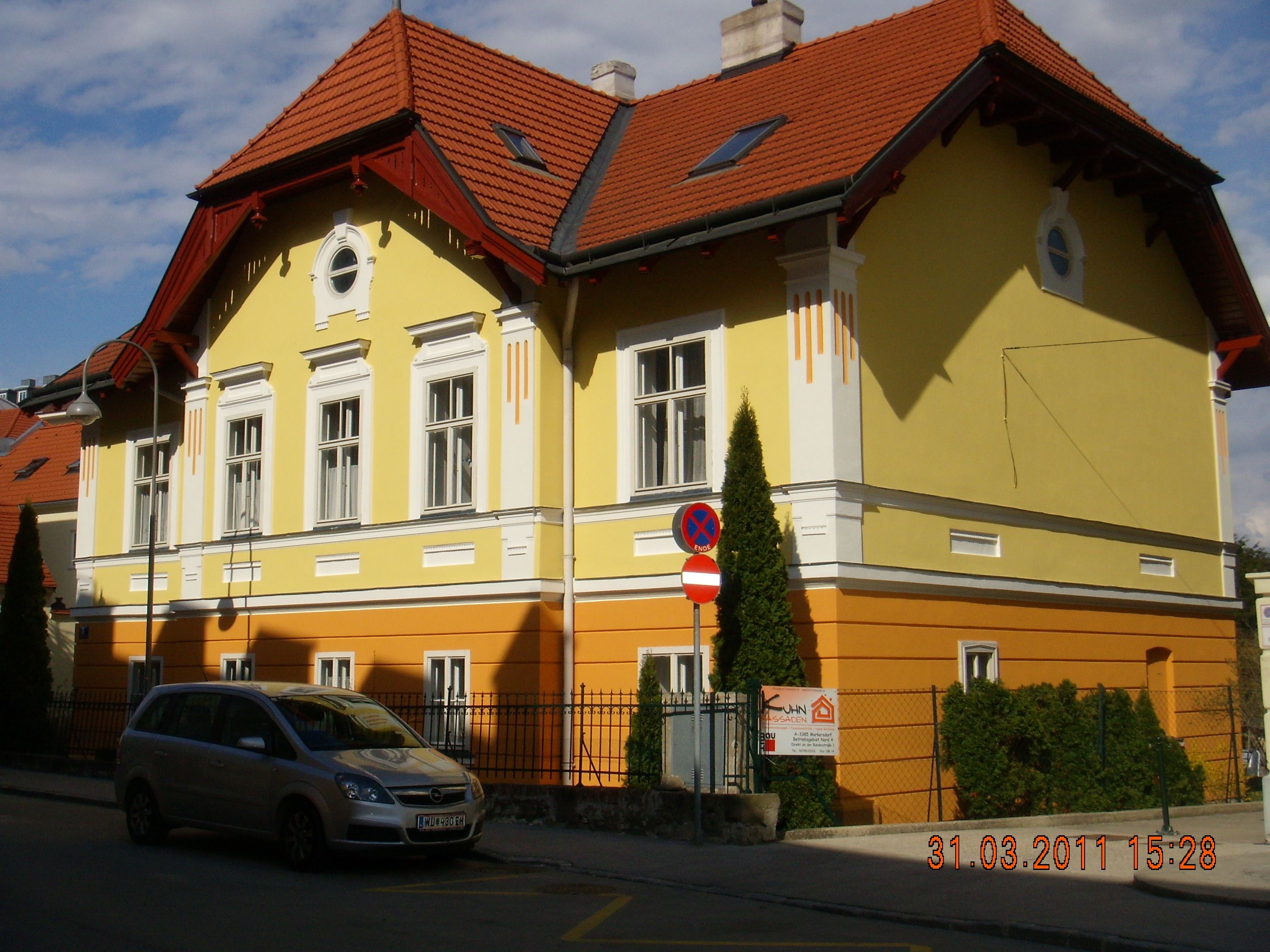 Gelbes und oranges Gebäude mit rotem Dach und geparktem Auto davor.