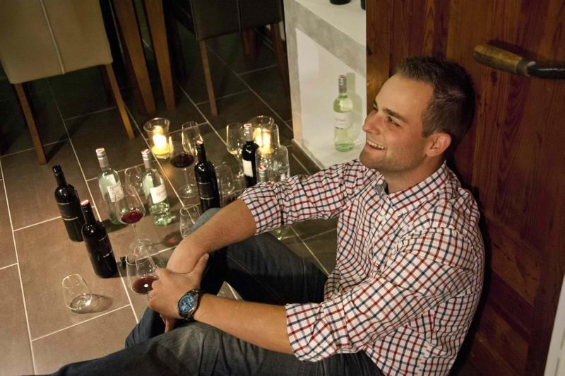 Ein Mann sitzt lächelnd auf dem Boden, umgeben von Weinflaschen und Gläsern.