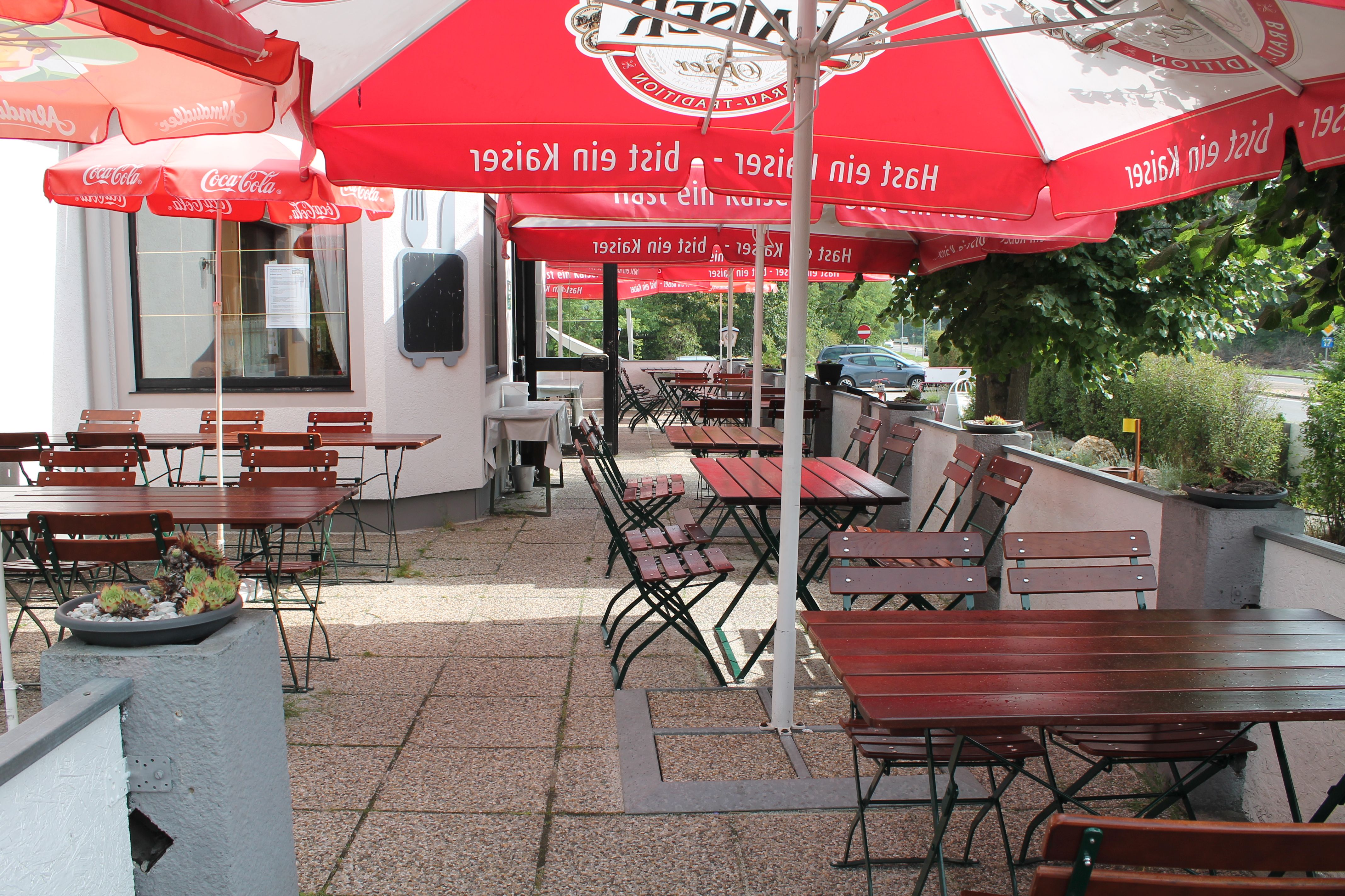 Ein leerer Gastgarten mit Holztischen und -stühlen unter roten Sonnenschirmen.