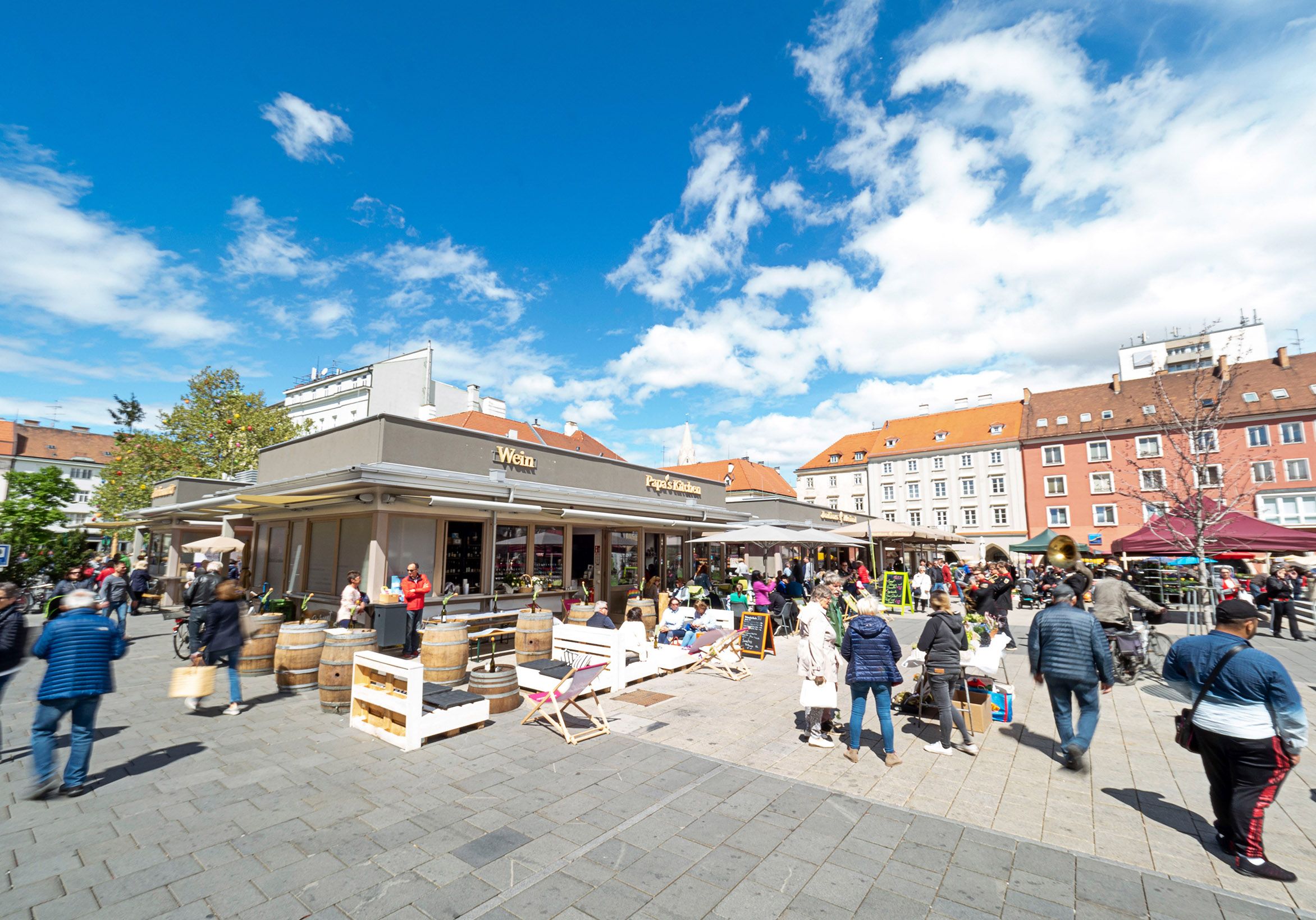 Ein belebter Marktplatz mit Menschen, die an Ständen und Cafés verweilen, unter einem blauen Himmel.