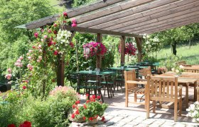 Ein gemütlicher Außenbereich eines Heurigen mit Holztischen und -stühlen, umgeben von blühenden Pflanzen und einem hölzernen Pergola-Dach.