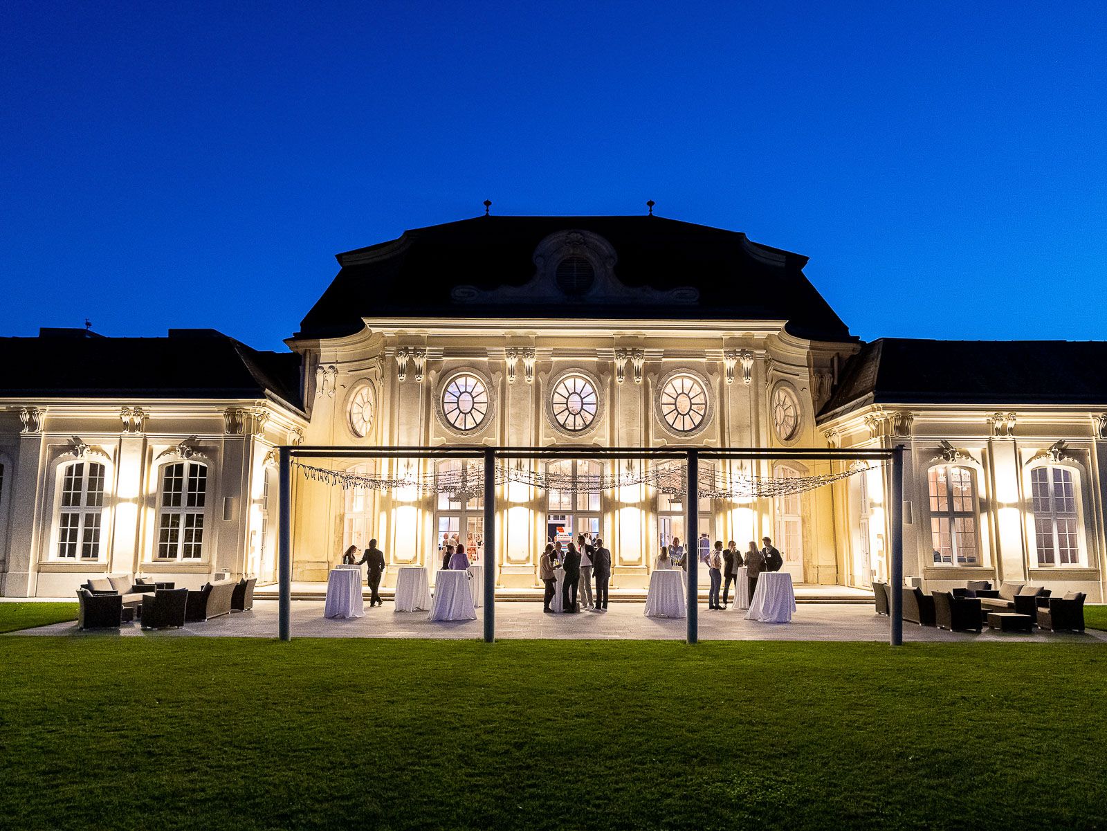 Conference Center Laxenburg bei Einbruch der Dunkelheit, Stehtische und einige Personen genießen das Ambiente.