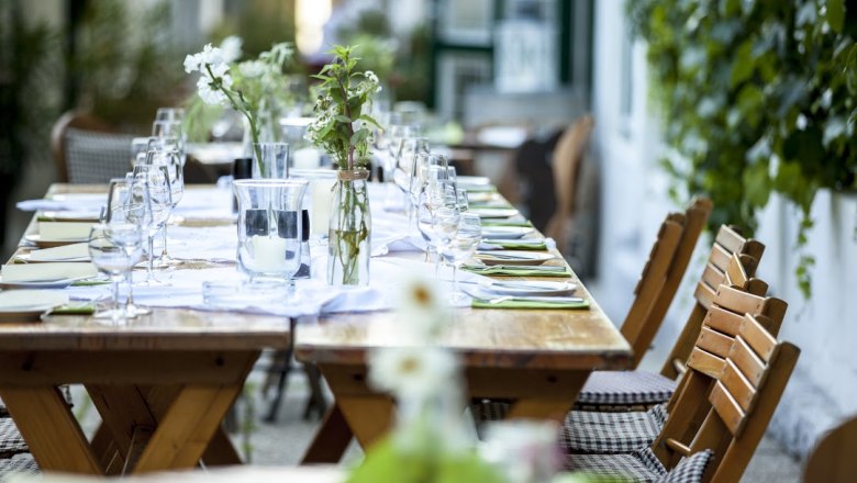 Langer Holztisch im Freien, gedeckt mit Gläsern, Besteck und Blumen.