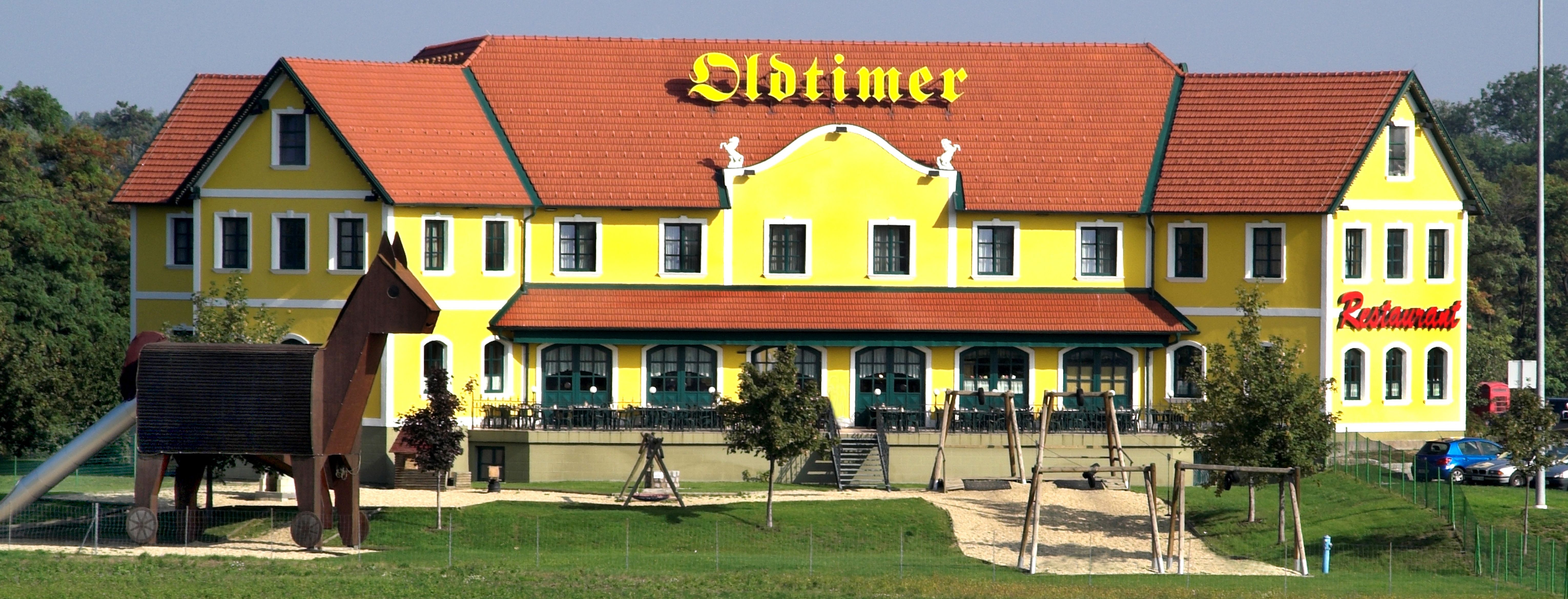 Gelbes Gebäude mit rotem Dach und der Aufschrift 'Oldtimer', daneben ein Spielplatz mit einer großen Holzfigur.