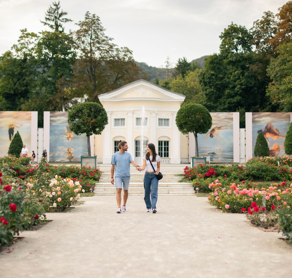 Zwischen blühenden Rosen im Doblhoffpark Baden spazieren eine Frau und ein Mann, vertieft in ein Gespräch. Im Hintergrund erhebt sich die eindrucksvolle Fotoausstellung von La Gacilly Baden Photo Baden, deren große Bilder das Szenario bereichern.
