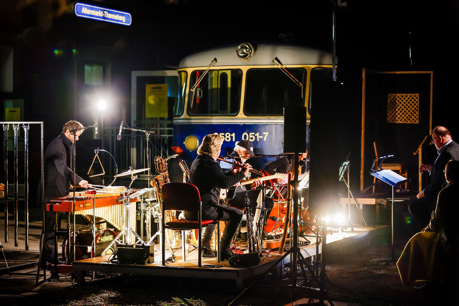 In der nächtlichen Kulisse des Kulturbahnhofs Altenmarkt entfaltet sich eine magische Atmosphäre, während Musiker harmonische Klänge erzeugen. Die sanften Lichter und die Präsenz der historischen Lokomotive schaffen einen einzigartigen Rahmen für ein unvergessliches Kulturereignis.
