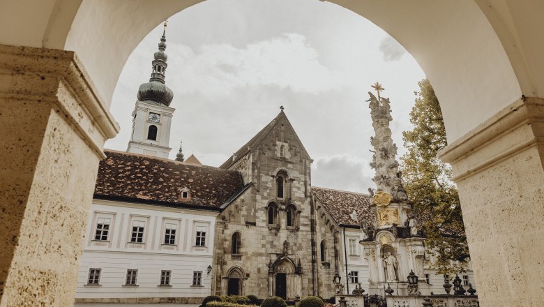 Blick durch einen Torbogen auf ein historisches Klostergebäude mit Turm und einer barocken Säule im Vordergrund.
