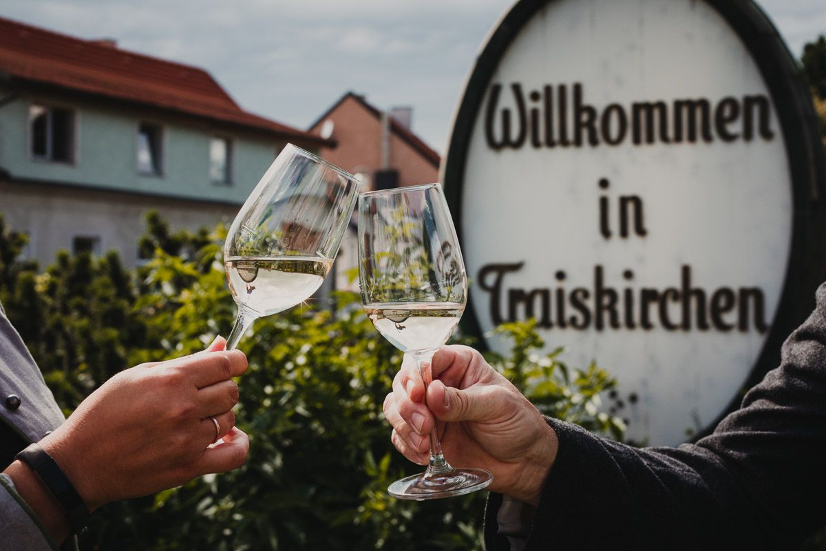 Im Ortskern von Traiskirchen steht ein Weinfass mit der Aufschrift Willkommen in Traiskirchen. Im Vordergrund sind zwei Hände sichtbar die mit einem Glas Weißwein anstoßen. 