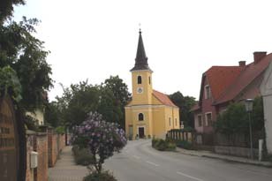 Eine gelbe Kirche mit Turm in einer ruhigen Straße, umgeben von Bäumen und Häusern.