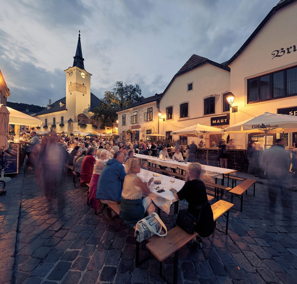 In der malerischen Kulisse des Wienerwaldes genießen die Gäste ein geselliges Beisammensein unter den schattigen Platanen. Der Duft von frisch gebackenem Brot und regionalen Weinen erfüllt die Luft und lädt zum Verweilen ein. Hier wird der Sommer in vollen Zügen gefeiert, während die sanften Hügel der Weinregion im Hintergrund leuchten.