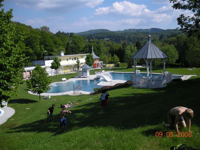 Ein Freibad mit Badegästen auf einer Wiese und einem Pavillon im Hintergrund.