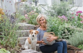 Eine Frau sitzt mit einem Hund in einem blühenden Garten auf Steinstufen.
