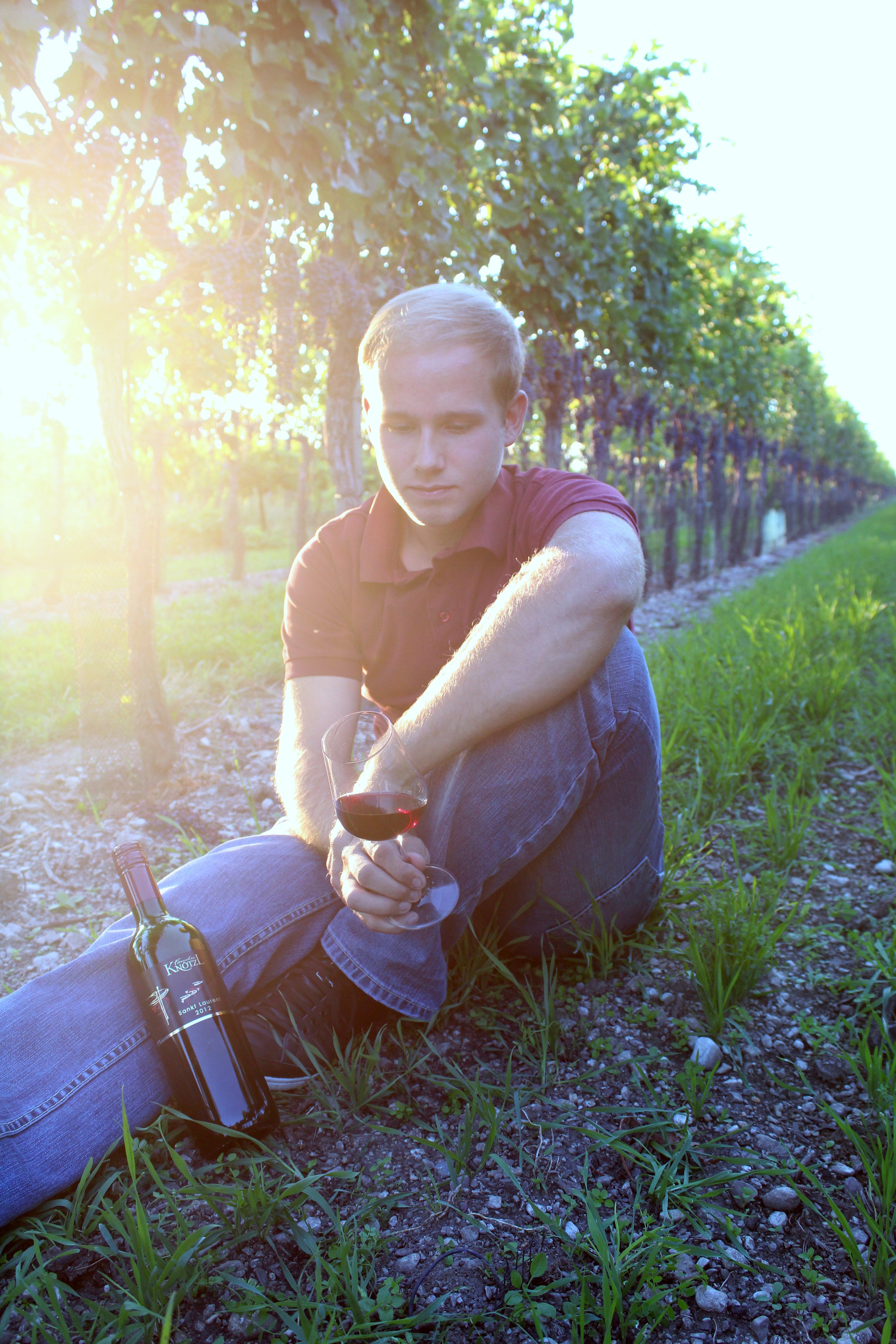 Ein junger Mann sitzt in einem Weinberg und hält ein Glas Rotwein. Neben ihm steht eine Weinflasche. Die Sonne scheint durch die Blätter.