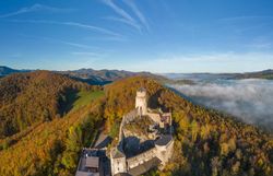 Luftaufnahme der Araburg im Herbst: Die Burg ist von orange- und gelb gefärbtem Wald umgeben, eine dichte Nebeldecke liegt über dem Tal, während oberhalb der Burg strahlender Sonnenschein den Himmel erhellt.