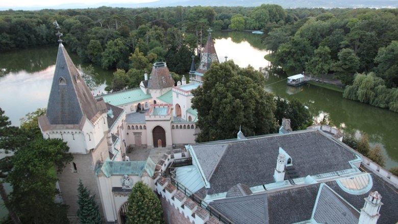 View of a castle with towers and a lake in the background, surrounded by trees.