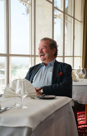 Frank Bläuel sitzt lächelnd im Restaurant des Berghotels Tulbingerkogel. Der Raum ist lichtdurchflutet, mit schön gedeckten Tischen, die der Umgebung eine warme und einladende Atmosphäre verleihen.