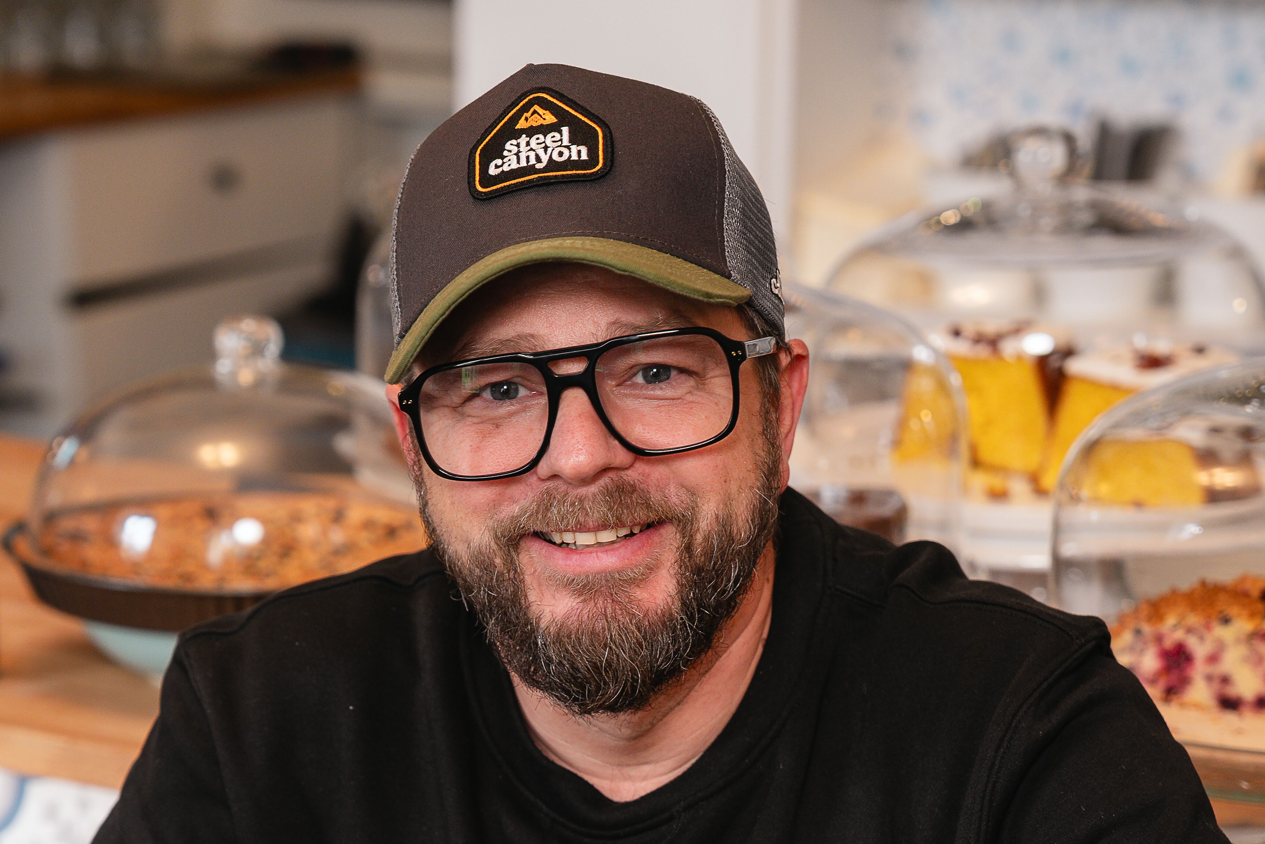 Michael Kulhanek sitzt entspannt in seinem Lokal "Kulhanek's Kaffee & Laden". Eine Reihe köstlicher Torten schmückt dahinter die Theke . Ein Ort, wo Genuss im Licht badet und die süßen Versuchungen voller Erwartung strahlen.