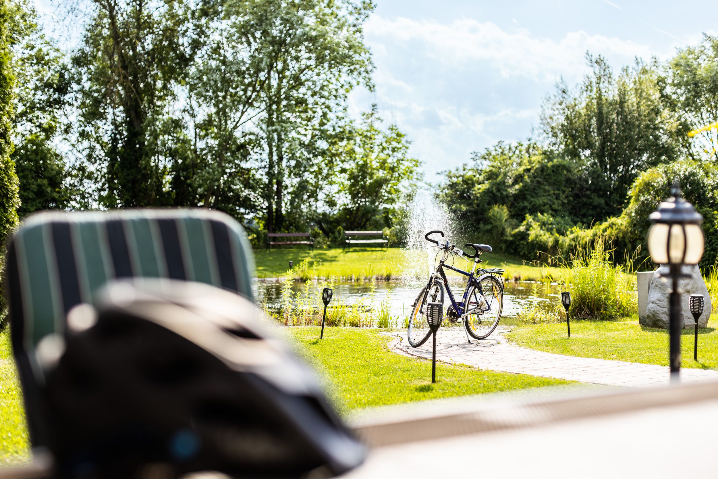 Ein Fahrrad steht neben einem Teich mit Springbrunnen in einem sonnigen Garten.