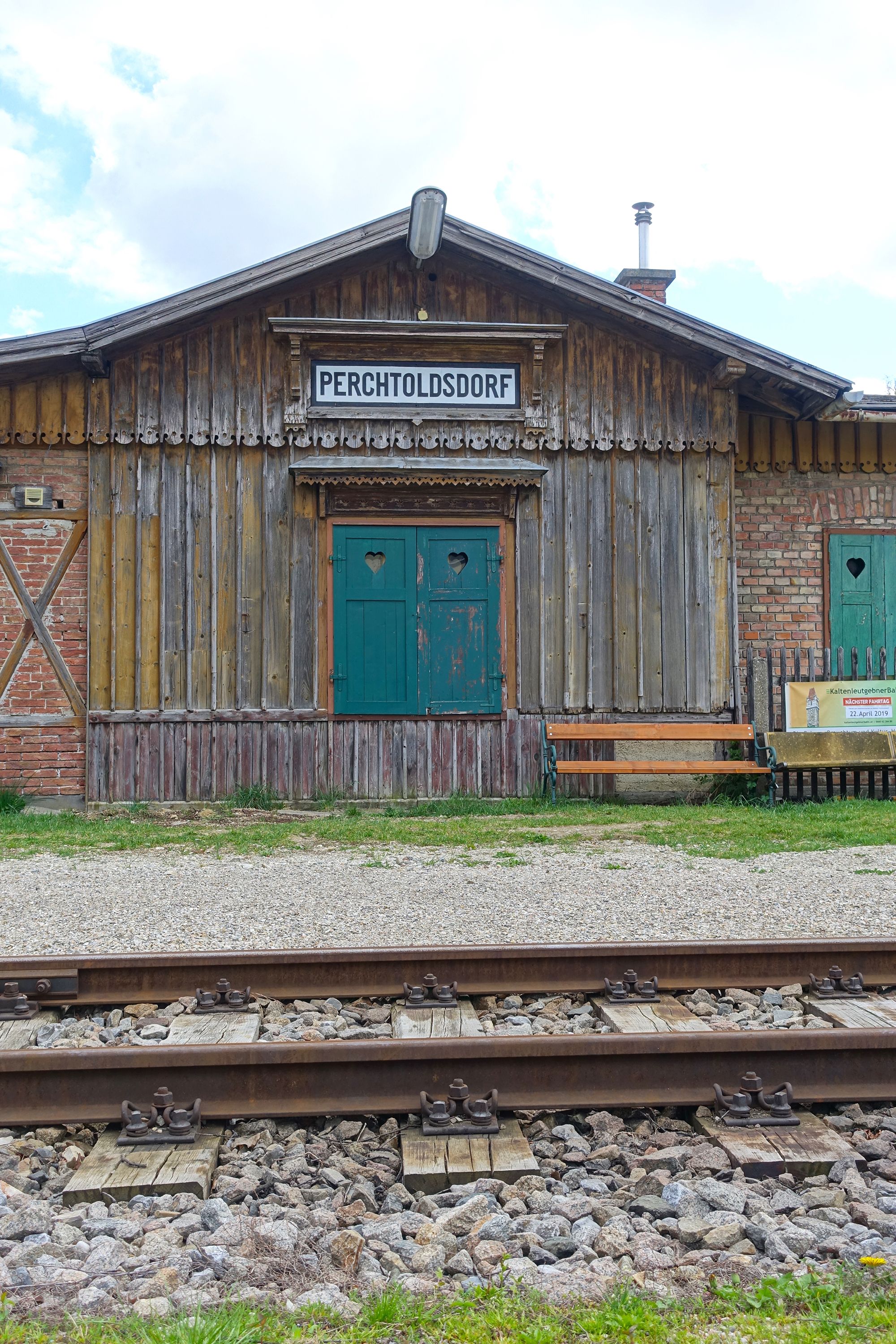 Versteckt zwischen den Zeilen der Vergangenheit steht ein hölzernes Bahnhofsgebäude in Perchtoldsdorf. Das Satteldach schützt, was von einer vergangenen Reisekultur geblieben ist – schlicht und doch eindrucksvoll. Ein einfaches Schild verkündet den Namen des Ortes und erinnert an Zeiten, als die Ankunft eines Zuges das Leben in Schwung brachte. Fenster und Türen erzählen leise Geschichten der Menschen, die einst hier ein- und ausgingen. 