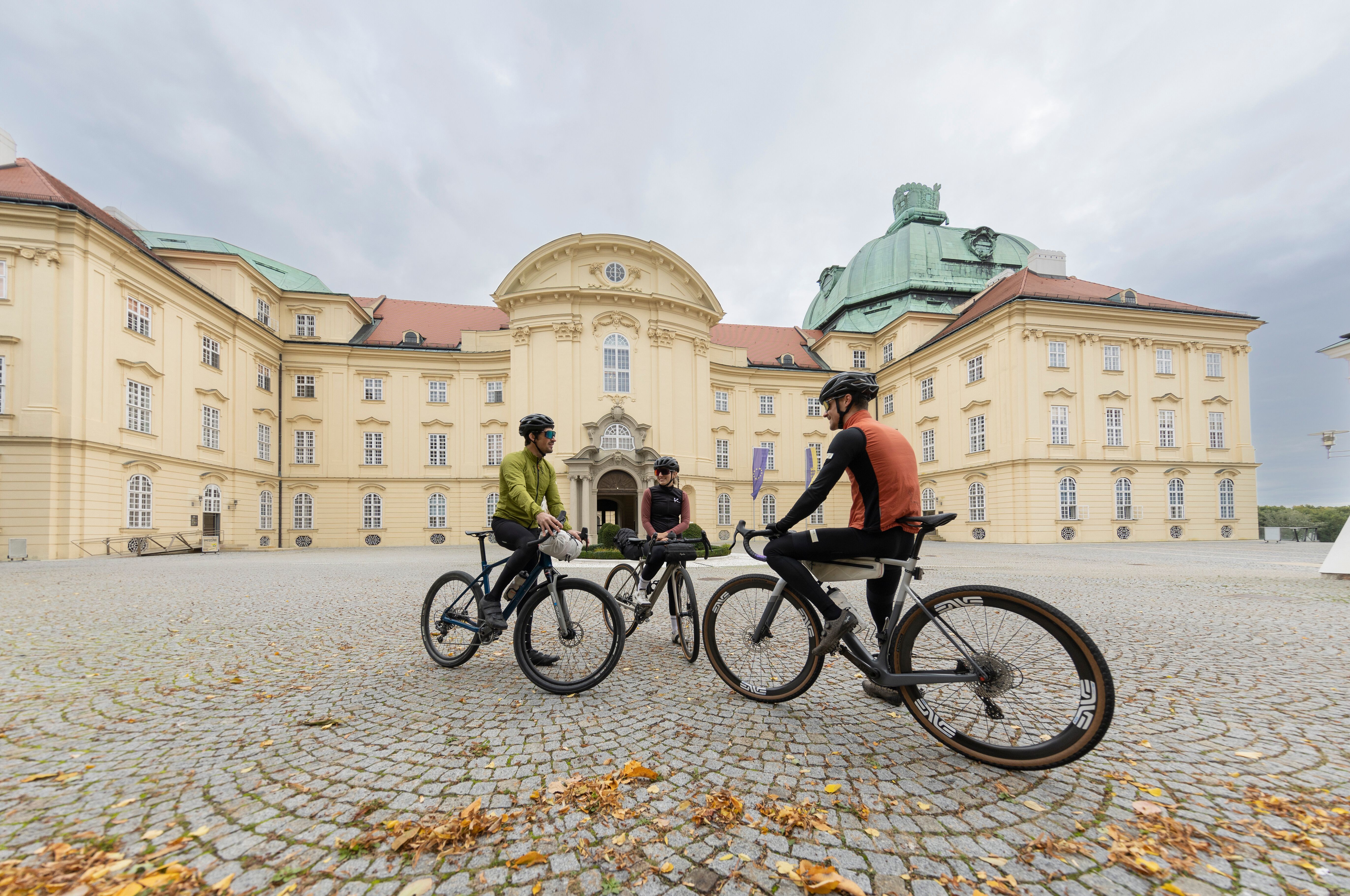 In der malerischen Kulisse des Wienerwaldes versammeln sich Radfahrer, um eine wohlverdiente Pause einzulegen. Die beeindruckende Architektur des Klosters im Hintergrund und die frische Luft laden dazu ein, die Schönheit der Natur zu genießen und neue Energie für die nächste Etappe zu tanken.