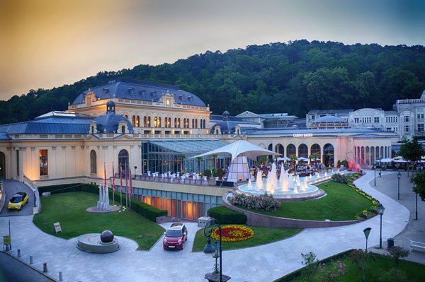 Außenansicht des Casino Baden mit beleuchteten Springbrunnen und umliegenden Grünflächen.