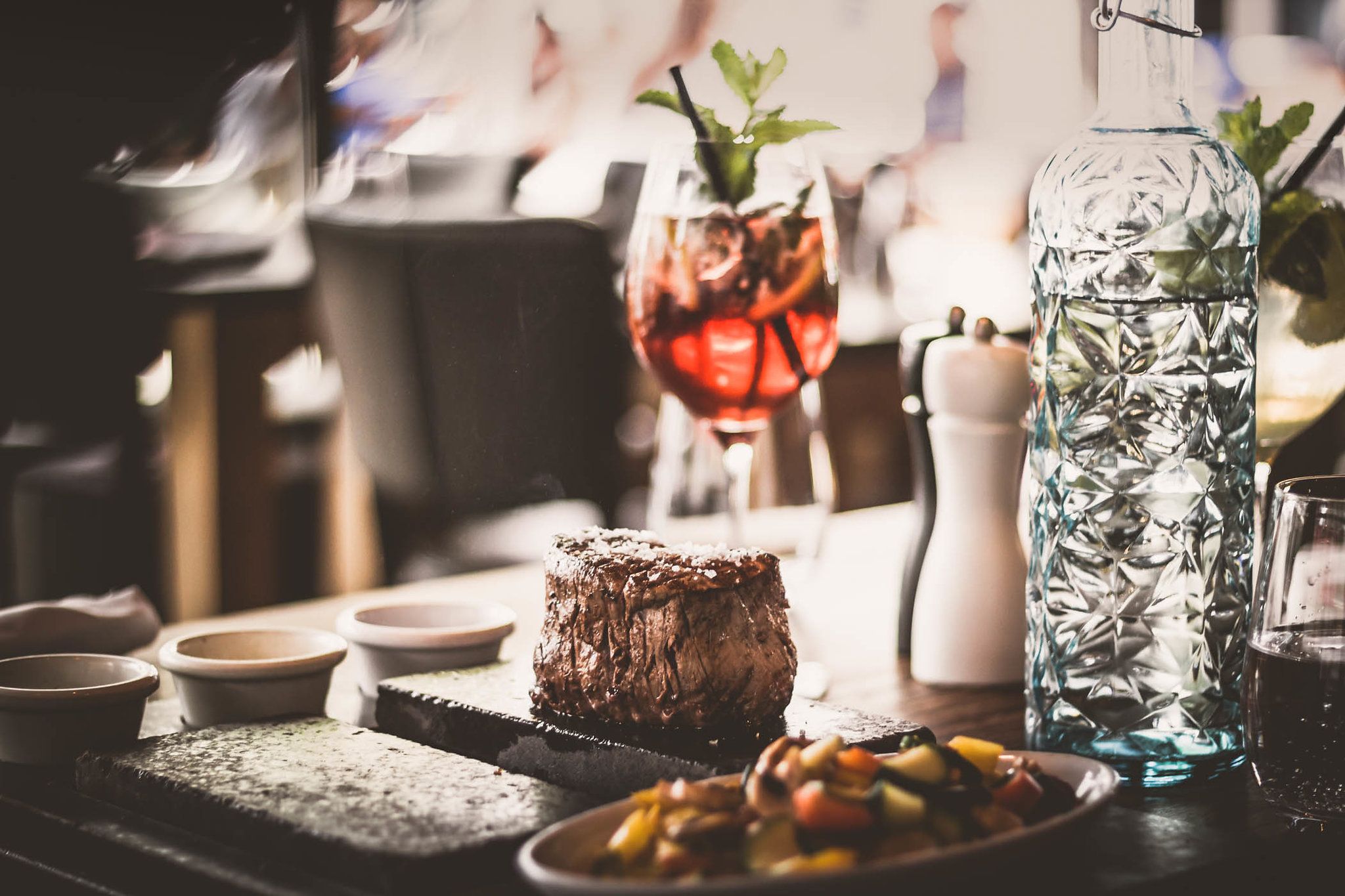 Ein saftiges Steak auf einem heißen Stein serviert, begleitet von Gemüse, einer Karaffe Wasser und einem Cocktailglas.