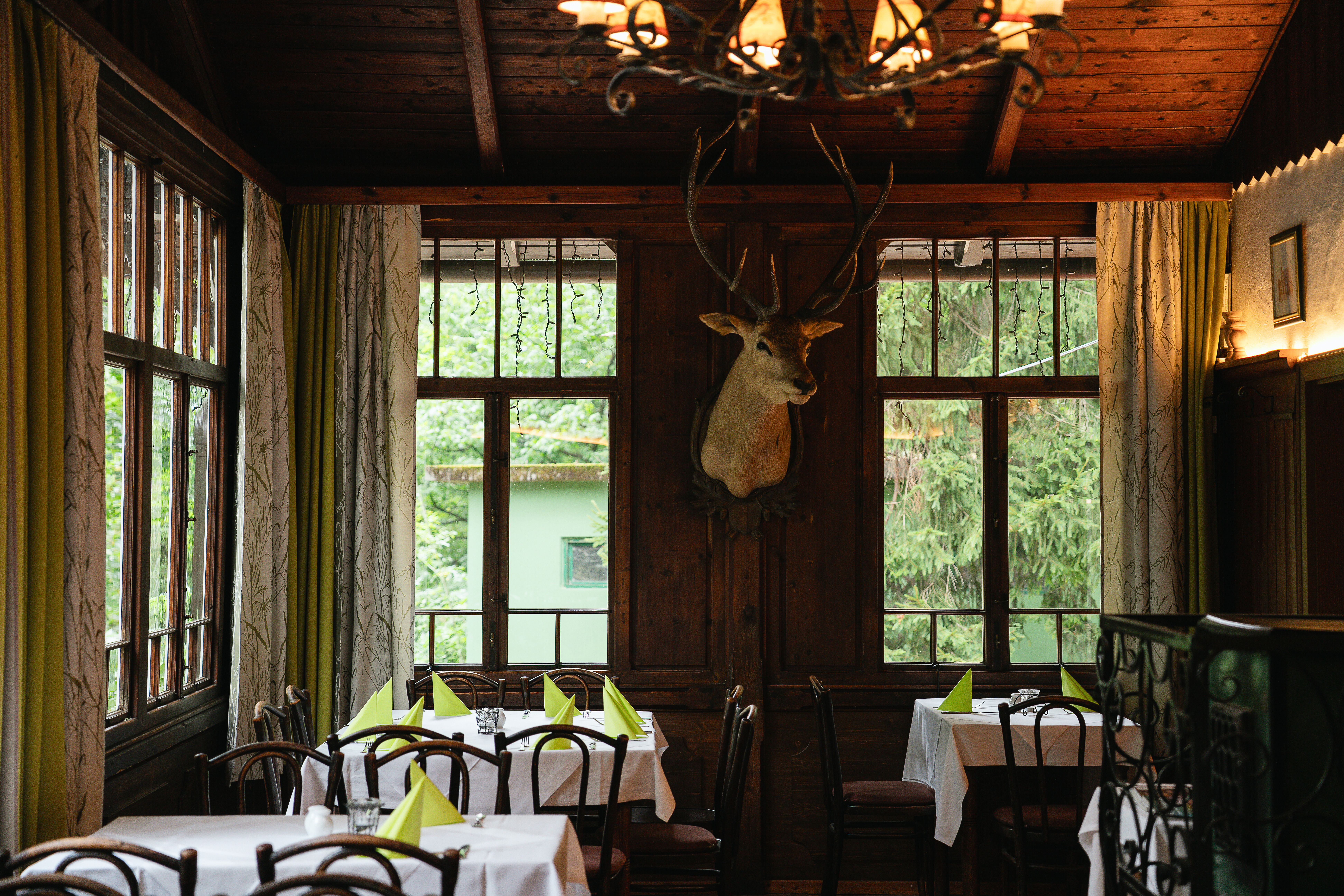 Im gemütlichen Restaurantbereich stehen dunkle Holzmöbel im Kontrast zu den lichtdurchfluteten Fenstern. Die Tische sind mit weißen Decken und leuchtend grünen Servietten gedeckt – ein farblicher Akzent, der Raffinesse versprüht. Dieser Raum lädt dazu ein, Momente der kulinarischen Entspannung zu genießen, inspiriert von der traditionellen Eleganz und lebendigen Natur.