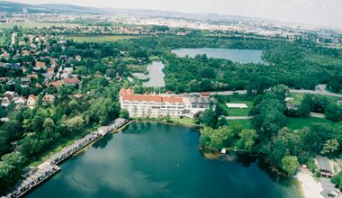 Luftaufnahme von Brunn am Gebirge mit einem großen Gebäude am See und grüner Landschaft.