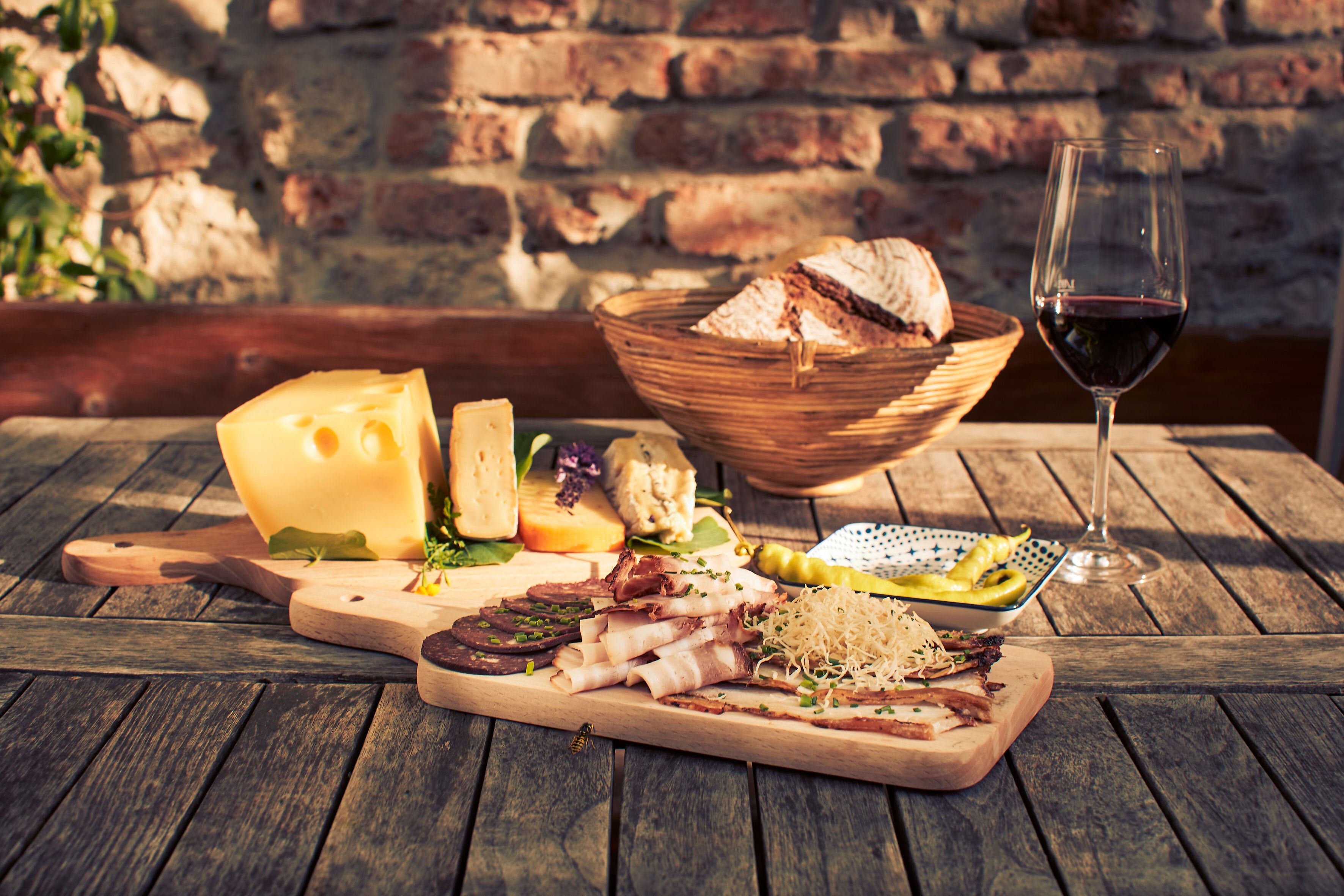 Genießen Sie eine köstliche Auswahl an regionalen Spezialitäten, während die warmen Sonnenstrahlen auf die rustikale Holzplatte scheinen. Ein Glas tiefroter Wein ergänzt die herzhaften Käsesorten und feinen Wurstwaren, die perfekt für einen entspannten Nachmittag im Freien sind.