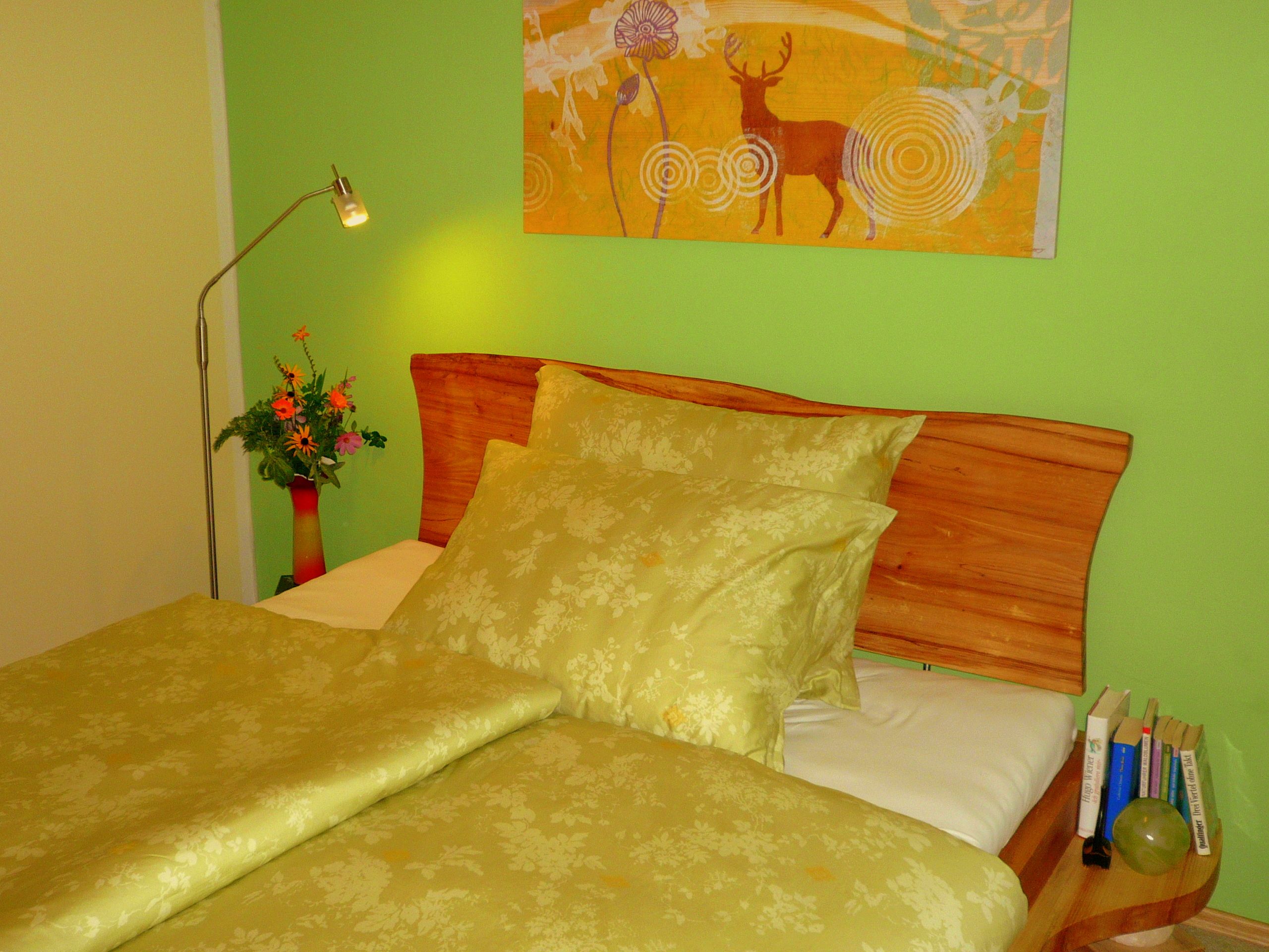 Gemütliches Schlafzimmer mit grünem Bettzeug, Holzkopfteil, grüner Wand und Hirschbild. Eine Lampe und Blumen stehen auf dem Nachttisch.