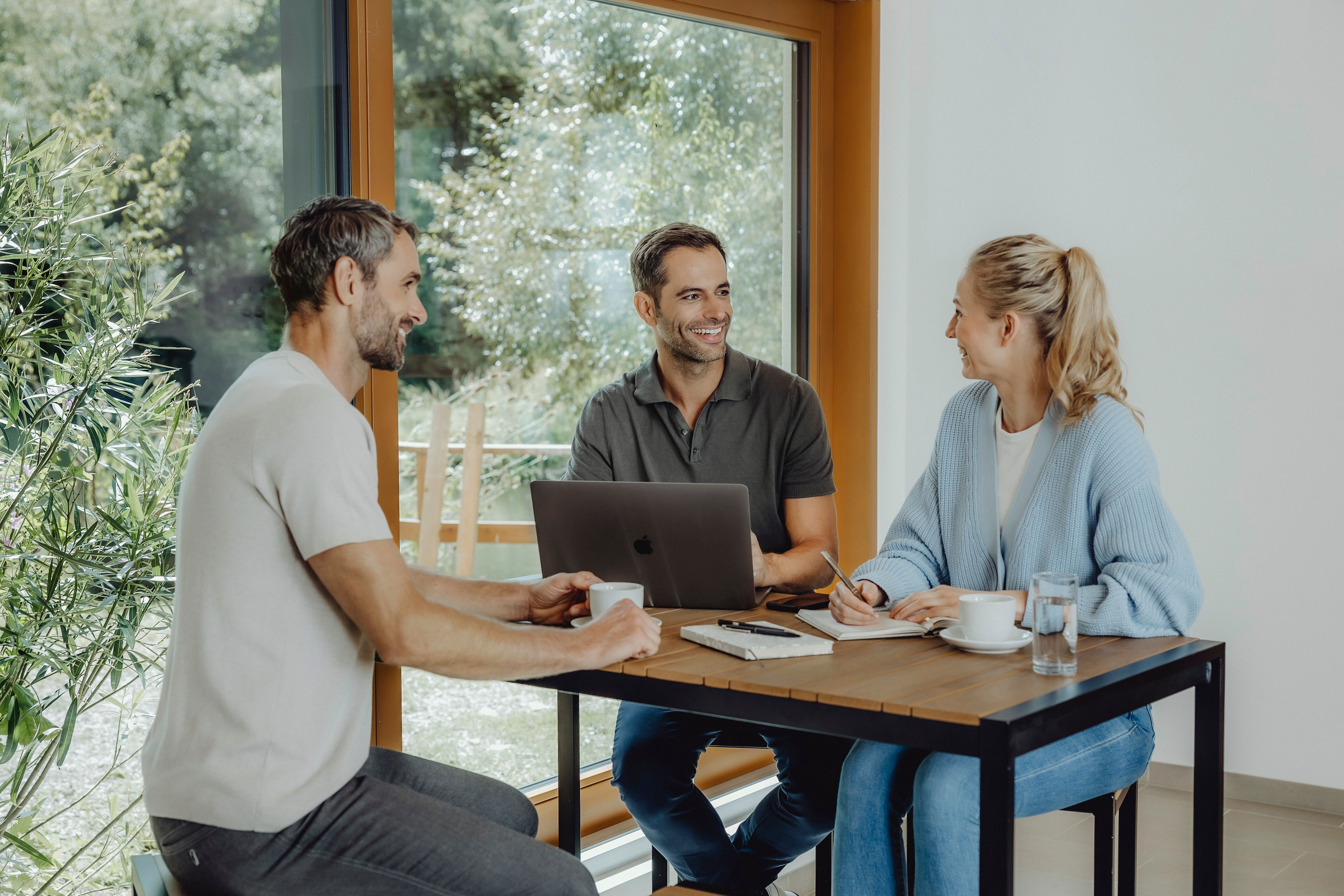 In einem hellen, einladenden Raum sitzen drei Personen um einen Tisch und genießen angeregte Gespräche. Die große Fensterfront lässt das Licht hereinströmen und bietet einen Blick auf die umliegende Natur, die zum Entspannen und Verweilen einlädt.