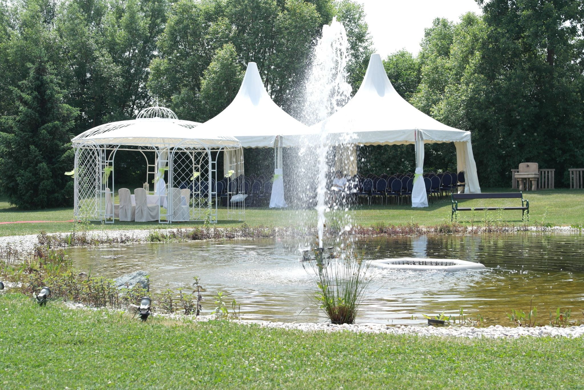Ein Hochzeitspavillon und Zelte neben einem Teich mit Springbrunnen in einem Garten.
