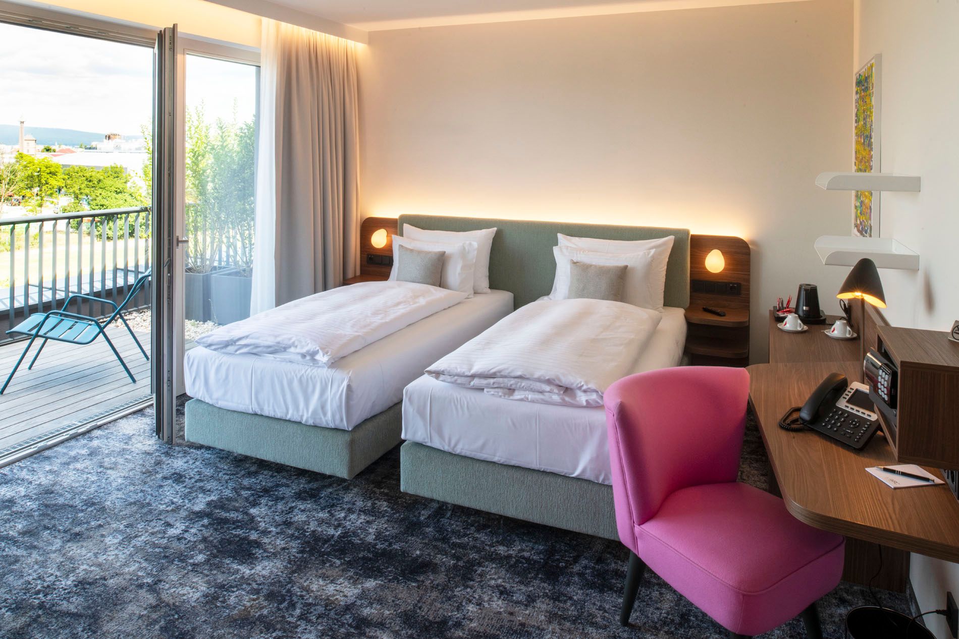 Modernes Hotelzimmer mit zwei Einzelbetten, einem Schreibtisch mit rosa Stuhl und Zugang zu einem Balkon mit Blick ins Grüne.