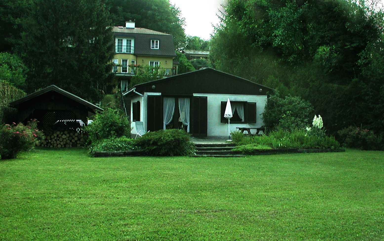 Ein kleines Haus mit Garten und Terrasse, umgeben von Bäumen und einem größeren Gebäude im Hintergrund.