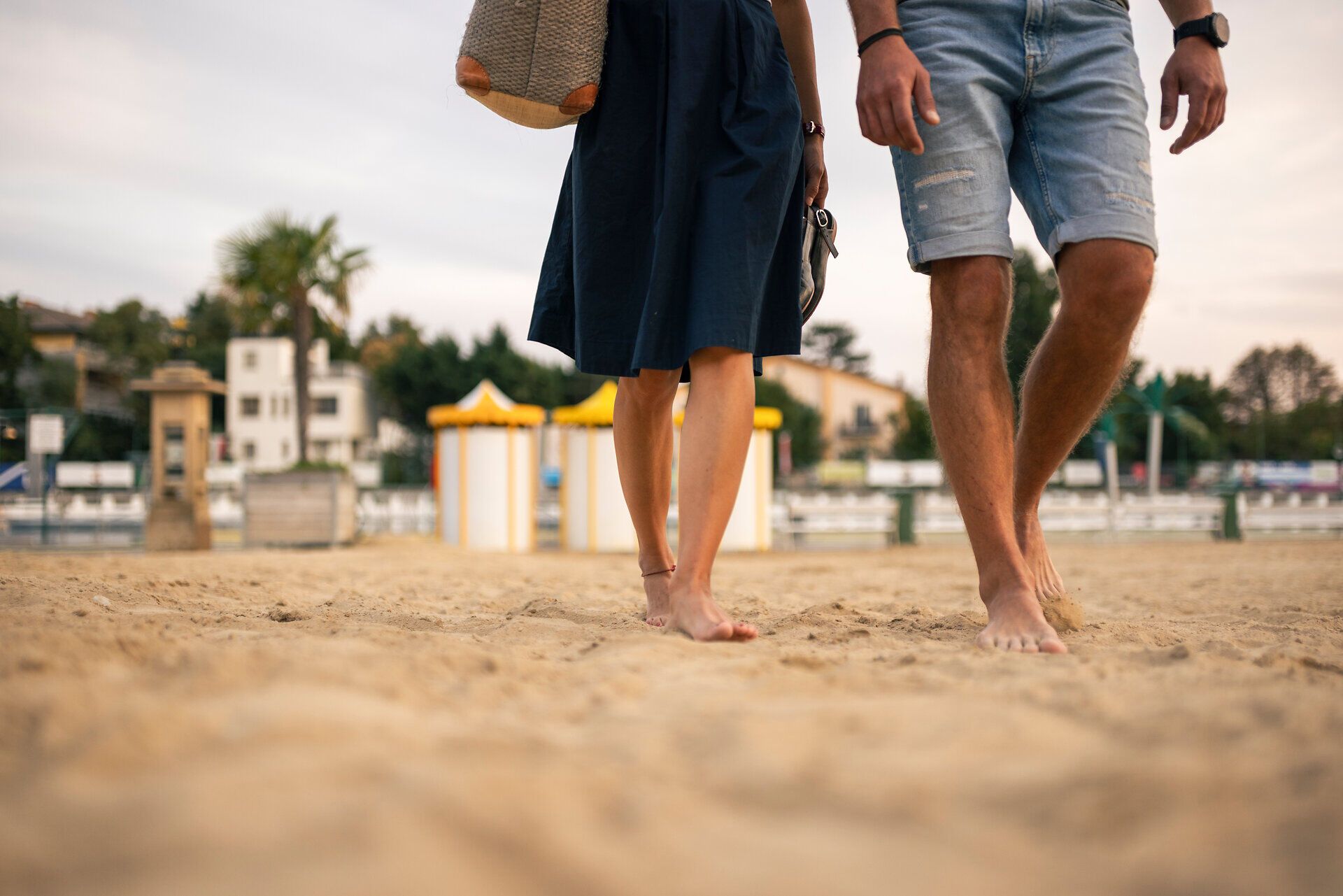 Ein Paar streift barfuß durch den Sand im Thermalstrandbad Baden, die Schuhe schwingen lässig in der Hand. Ein Augenblick voller Leichtigkeit, der die Magie eines sommerlichen Abends im Wienerwald einfängt.