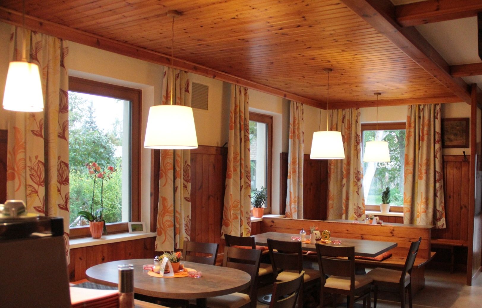 Gemütliche Gaststube mit Holzmöbeln, großen Fenstern und hängenden Lampen.
