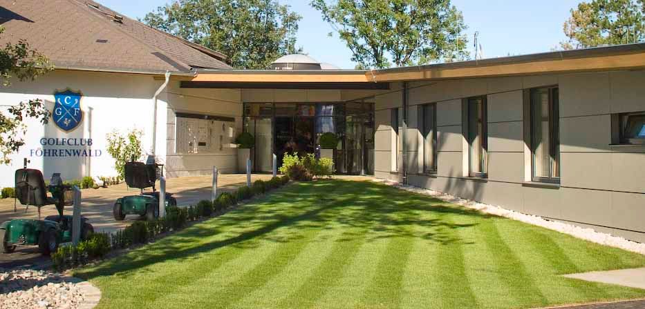 Eingang des Clubhauses des Golfclubs Föhrenwald mit gepflegtem Rasen und modernen Gebäuden.