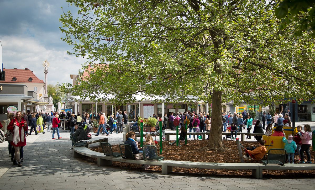 Menschenmenge auf einem Marktplatz mit Bäumen und Spielplatz.