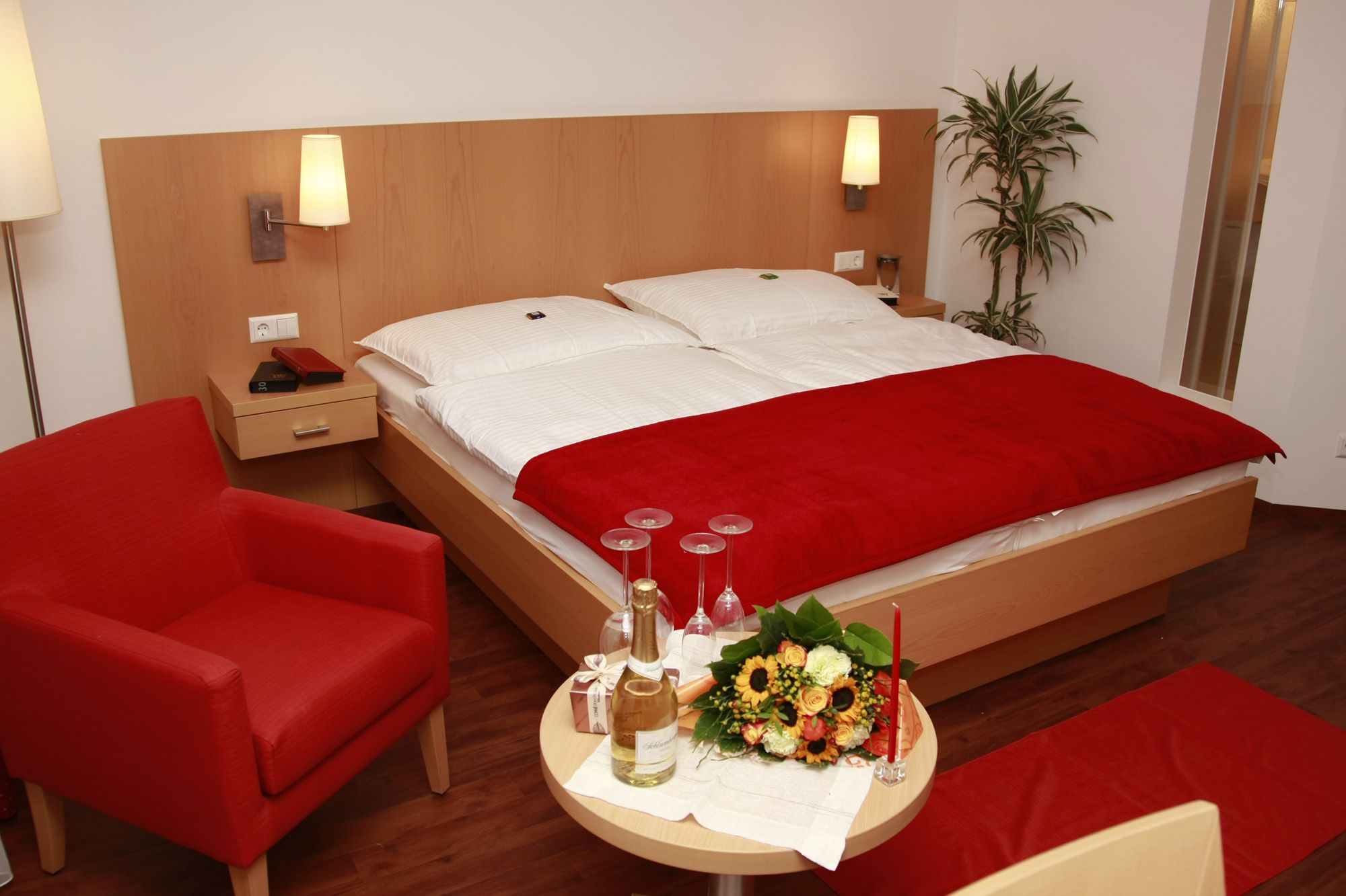 Ein gemütliches Doppelzimmer mit rotem Sessel, Blumenstrauß und Sekt auf einem Tisch.