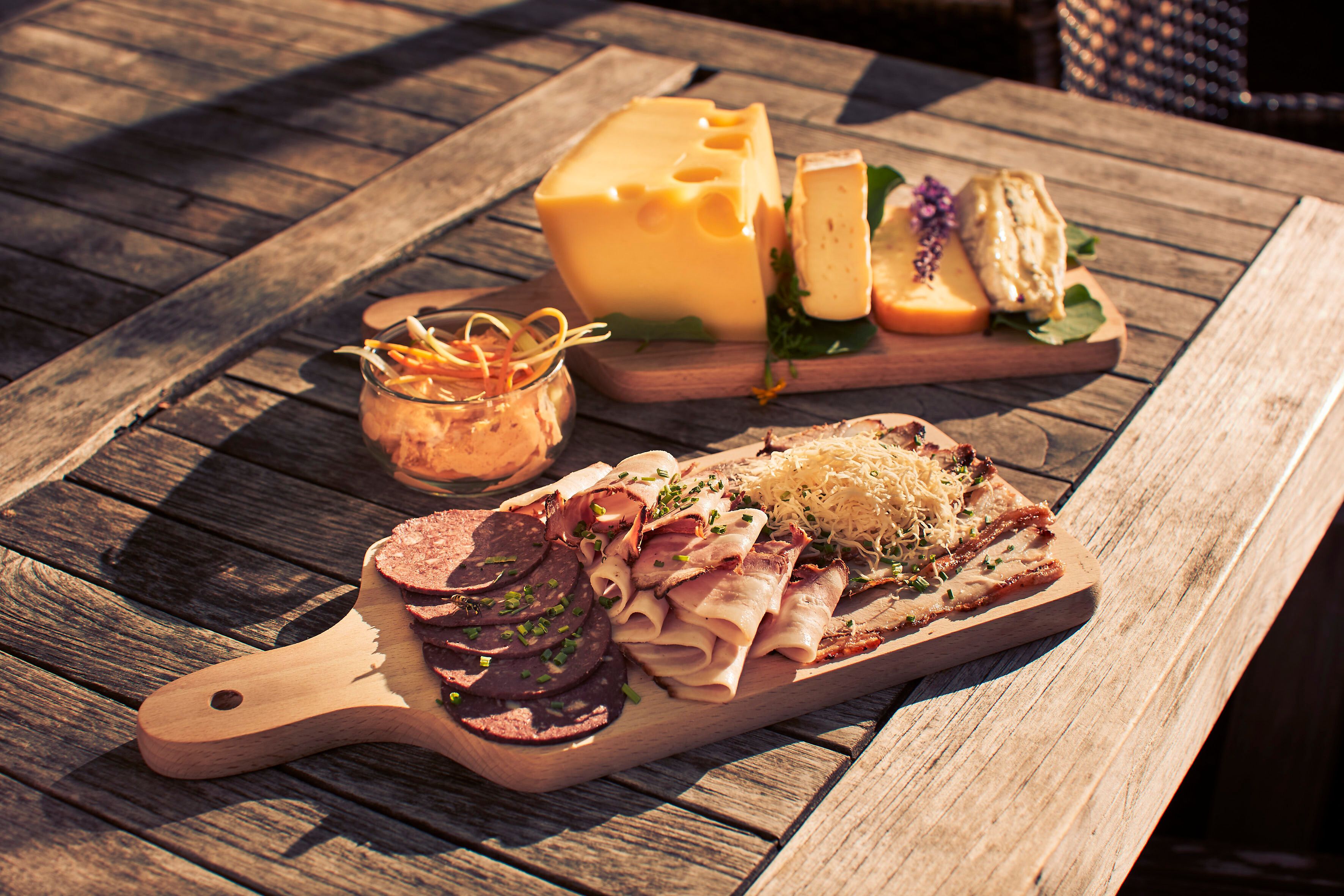 Genießen Sie eine köstliche Auswahl an regionalen Delikatessen, während die warmen Sonnenstrahlen auf die rustikale Holzterrasse scheinen. Die harmonische Kombination aus feinen Wurstwaren und aromatischen Käsesorten lädt dazu ein, die Seele baumeln zu lassen und die Schönheit der Umgebung zu erleben.