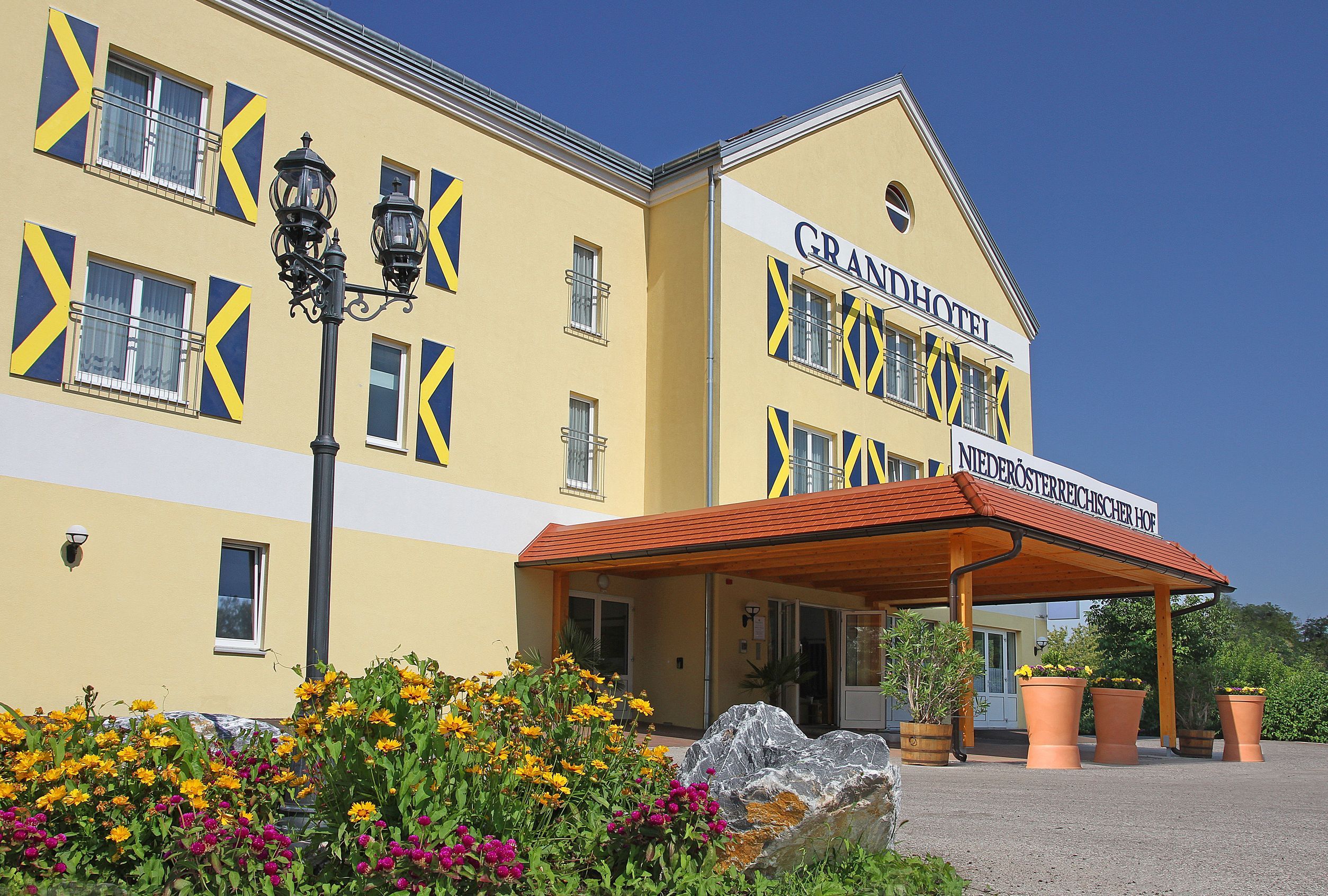 Außenansicht eines gelben Hotels mit blauen Fensterläden und einem Vordach. Vor dem Gebäude blühen gelbe und pinke Blumen.