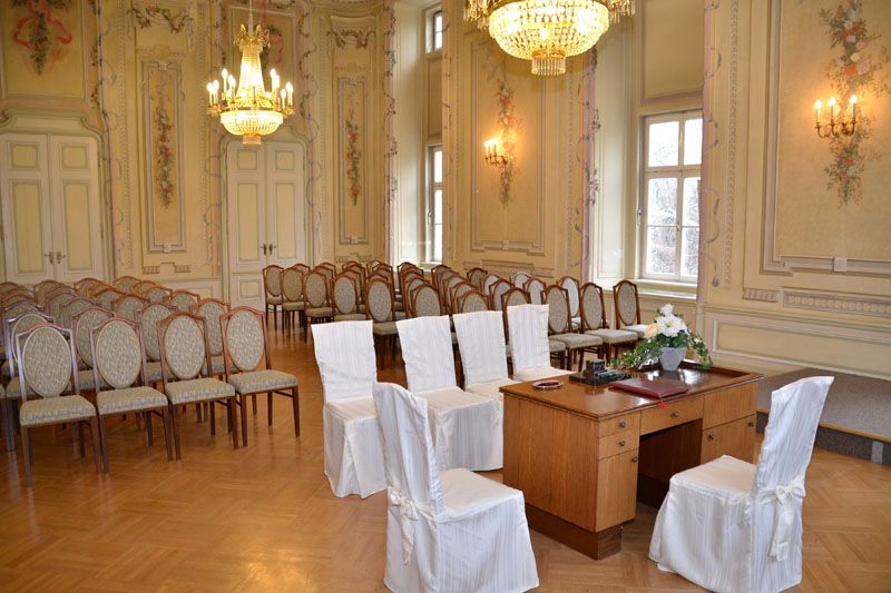 Eleganter Festsaal mit Stühlen, die mit weißen Hussen bezogen sind, und einem Holztisch. Kronleuchter und verzierte Wände schmücken den Raum.