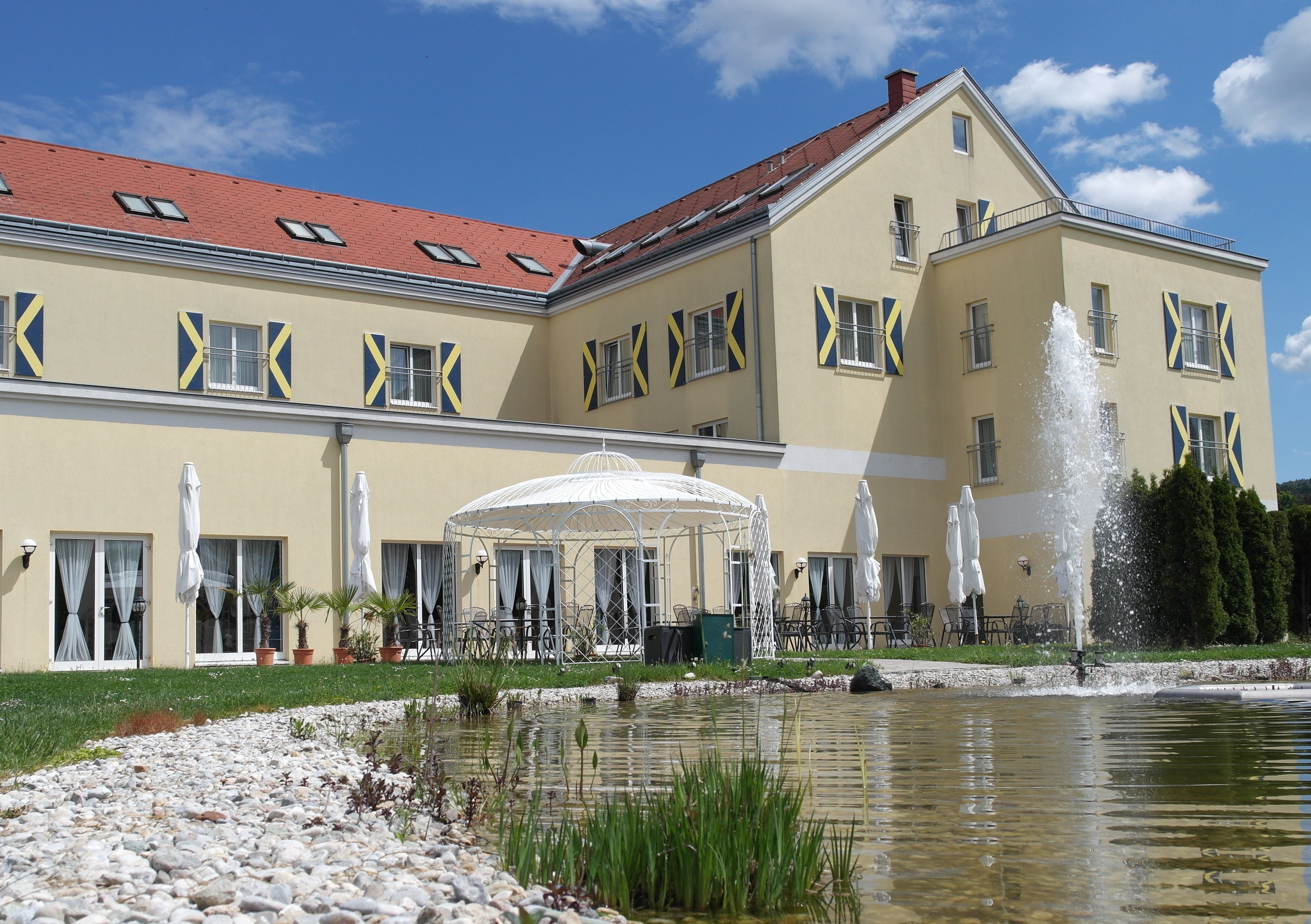 Großes gelbes Gebäude mit roten Dachziegeln, Springbrunnen und Pavillon im Vordergrund.
