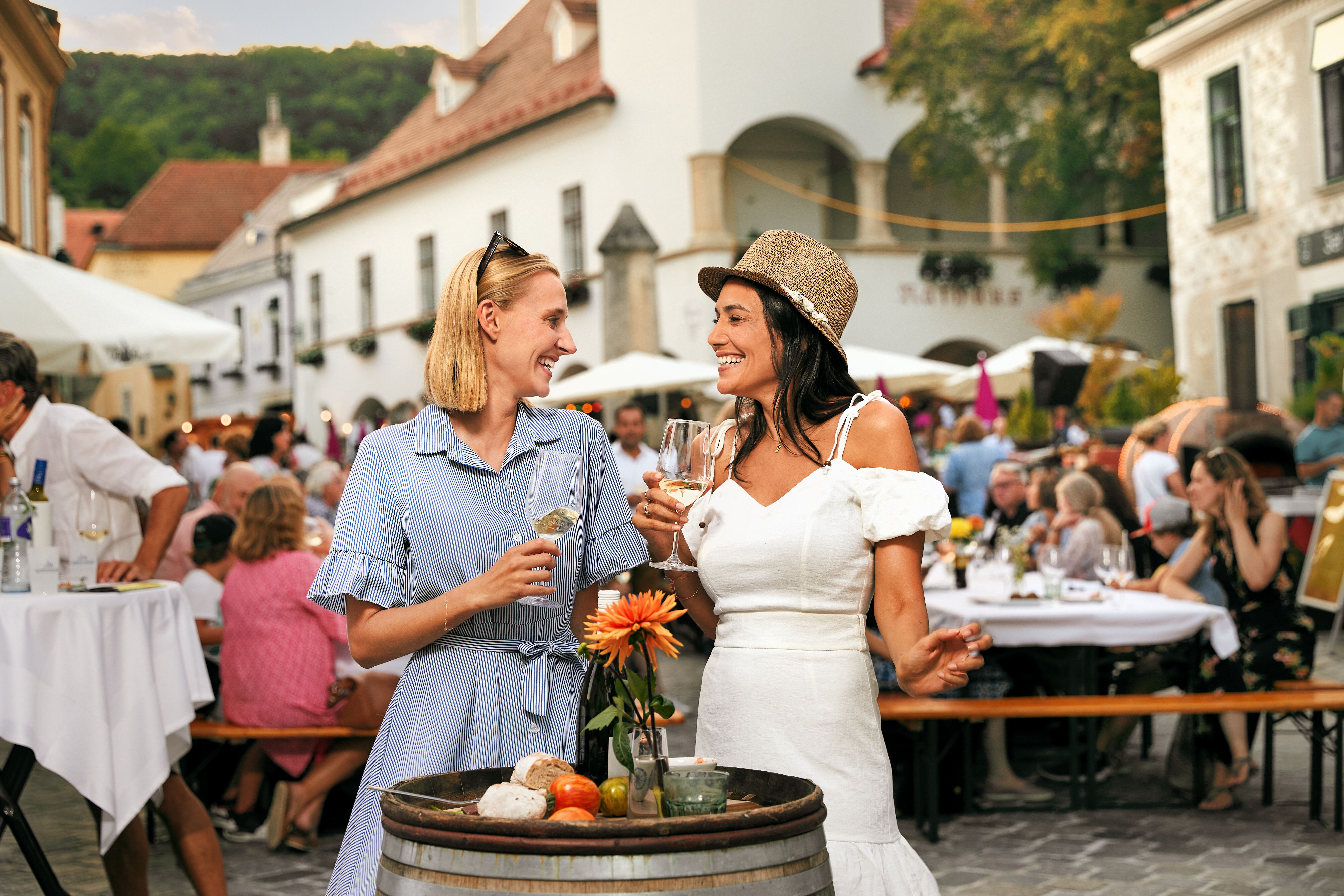 Zwei Frauen genießen ein fröhliches Gespräch bei einem Weinfest, umgeben von festlicher Atmosphäre und kulinarischen Köstlichkeiten. Die warmen Sonnenstrahlen tauchen die Szenerie in ein goldenes Licht, während die Tische mit frischen Blumen und erlesenen Weinen geschmückt sind.