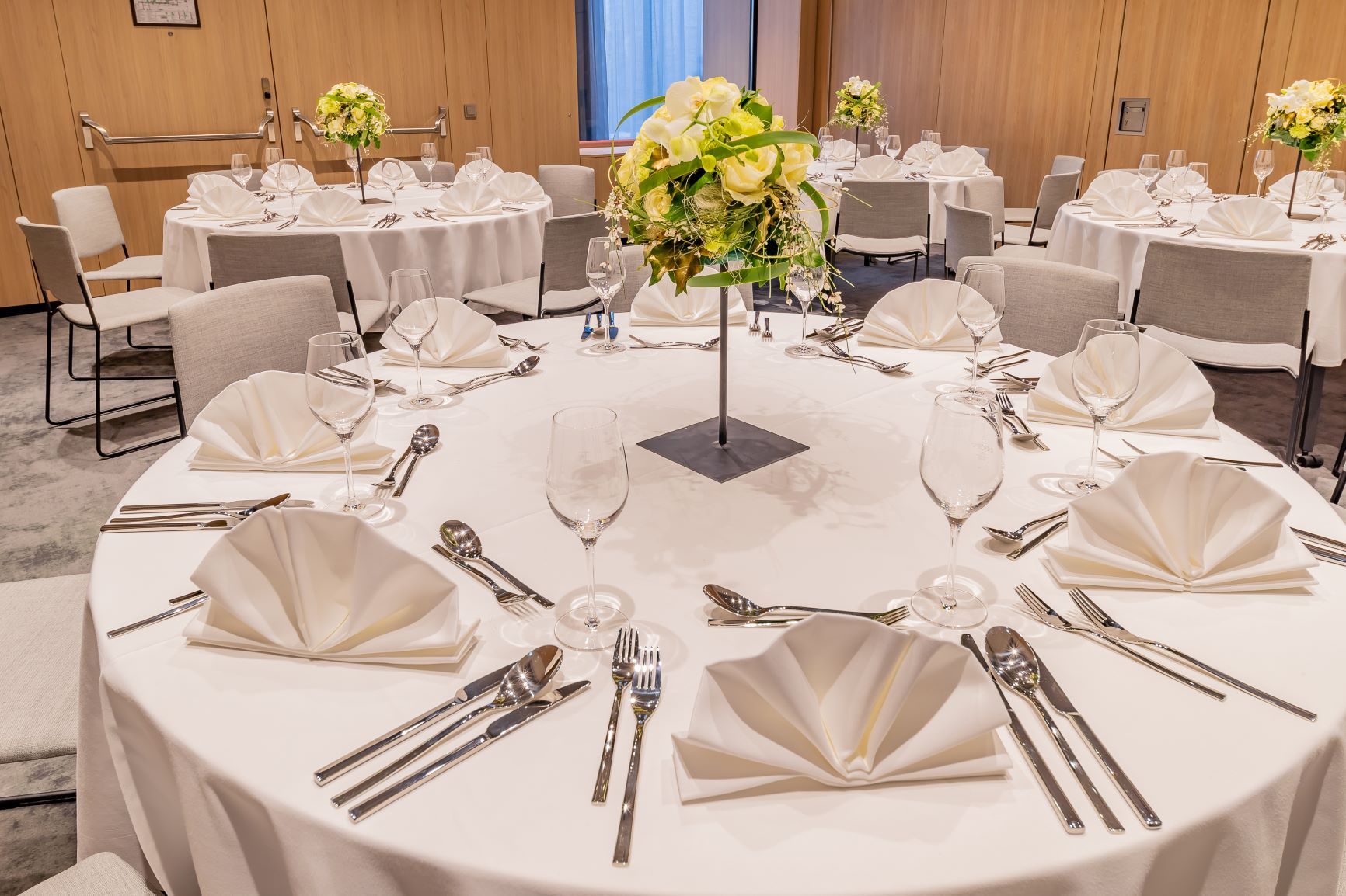 Rund gedeckter Tisch mit weißer Tischdecke, Besteck, Gläsern und Blumenarrangement in einem eleganten Raum.