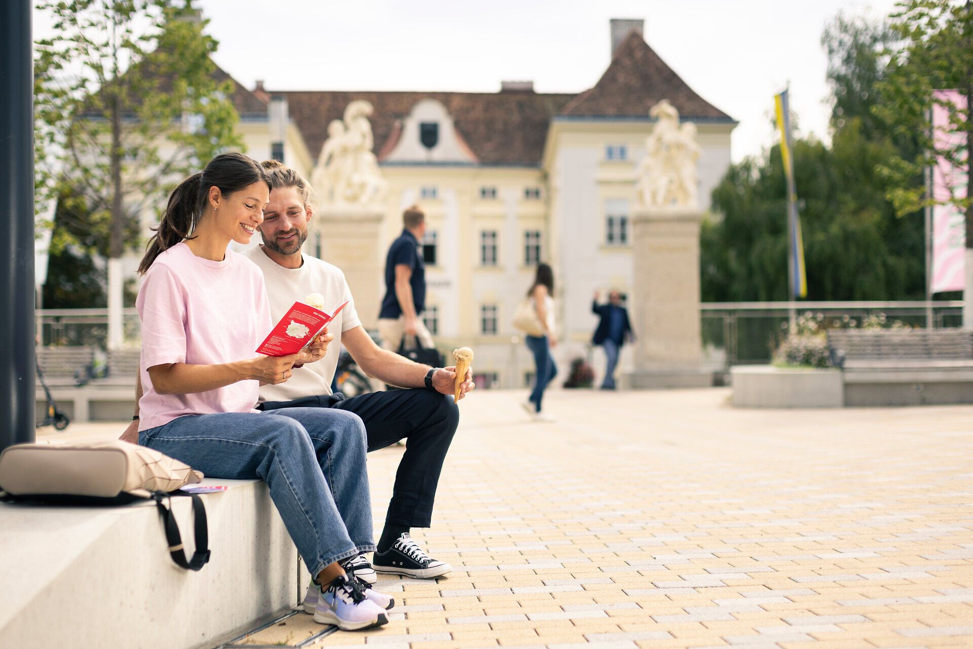 Ein Mann und eine Frau sitzen an einem Sommertag vor dem Schloss Bad Vöslau und studieren eine Broschüre. Im Hintergrund sind Personen zu sehen, die über den Schlossplatz schlendern.