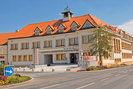 Rathaus Traiskirchen mit rotem Dach und mehreren Fenstern, umgeben von einer Straße und Blumenbeeten.