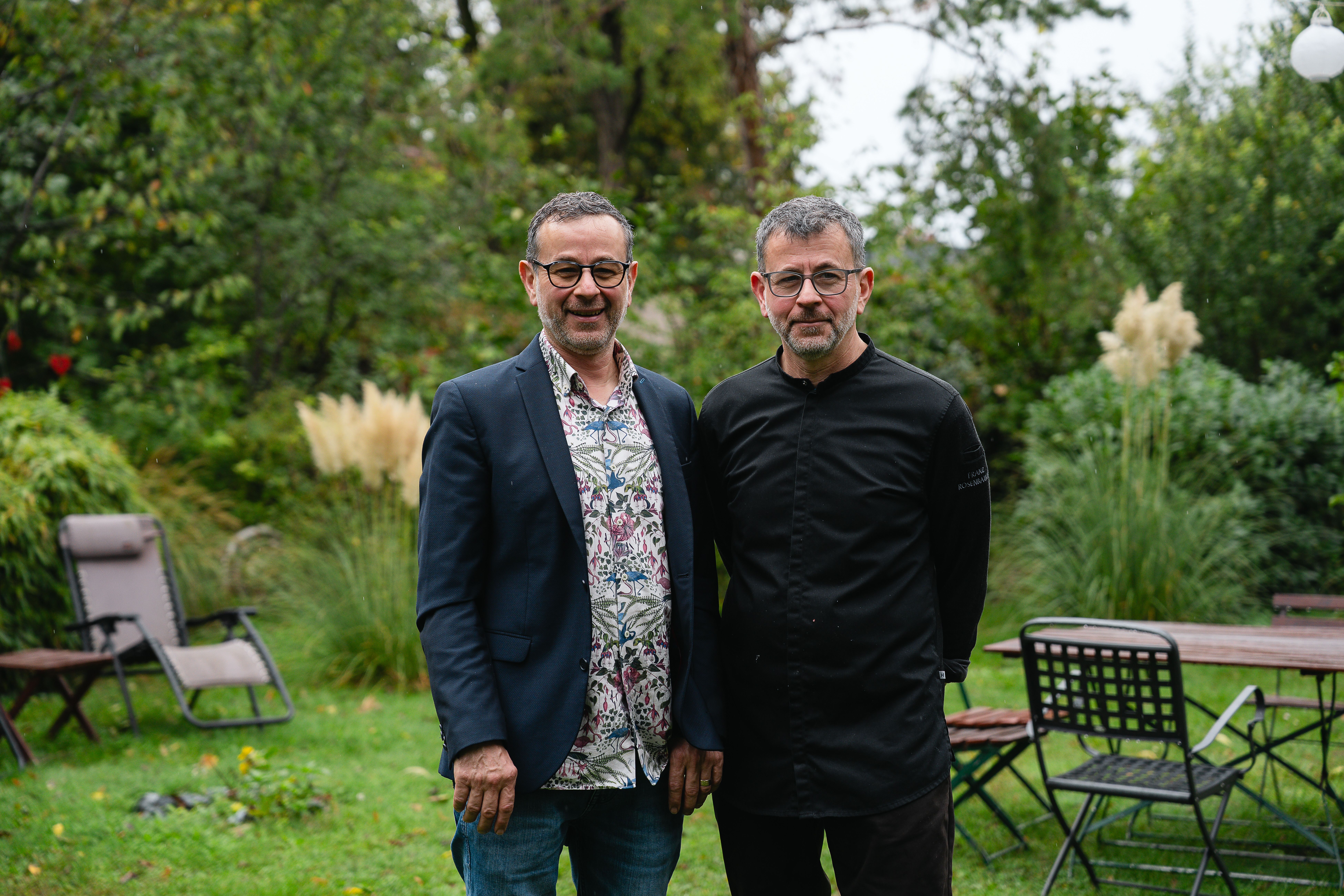 Die Brüder Franz und Karl Rosenbauch stehen nebeneinander in ihrem großen Garten, umgeben von Bäumen und Grün, beide lächelnd.